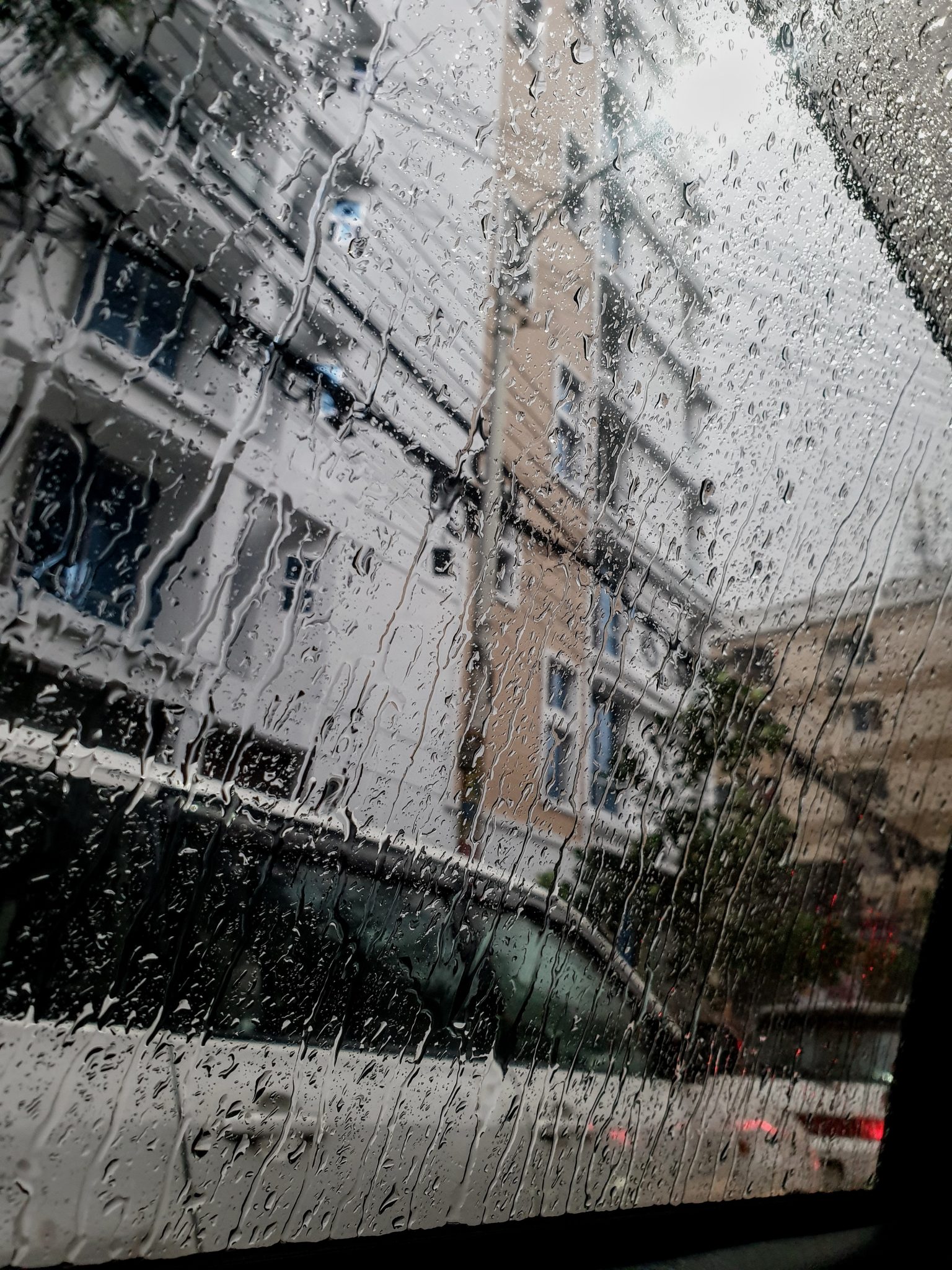 Raindrops on a car window blur the view of tall buildings and a parked white car outside. The scene 