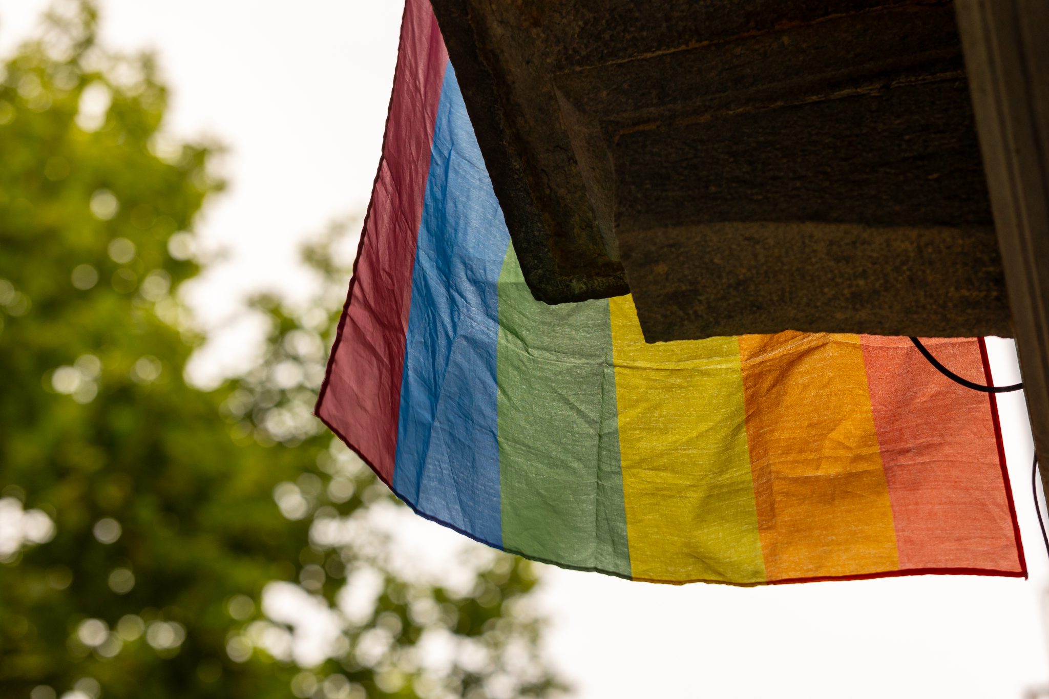 Bottom of a wrinkled pride flag hanging from a balcony with a tree in the background