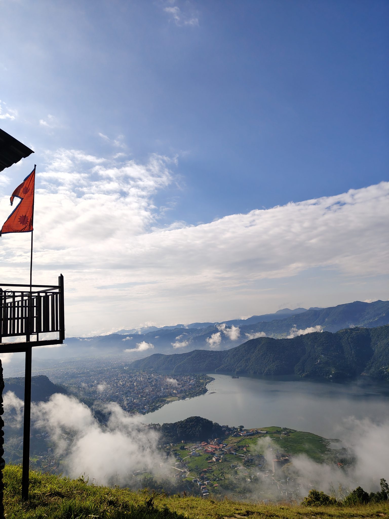From a wooden balcony with a red flag, a panoramic view spans Pokhara’s valley, Lake Phewa, the city
