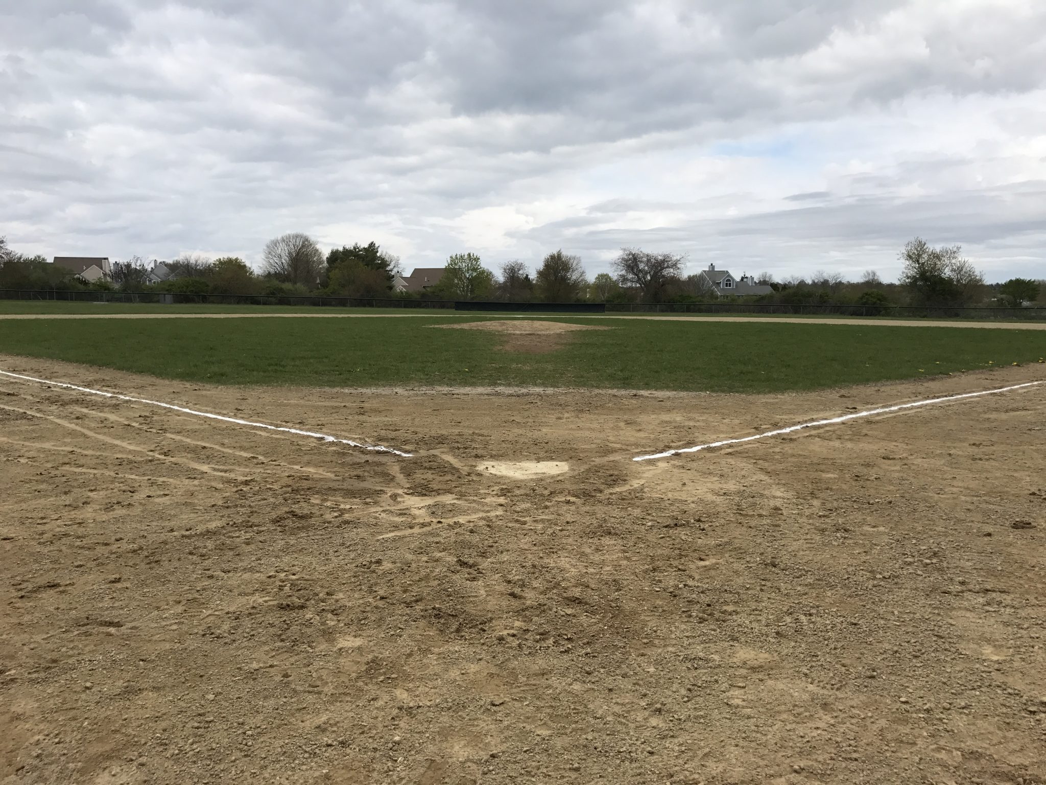 A baseball field with freshly chalked foul lines on an overcast day. Rooftops are visible over the t