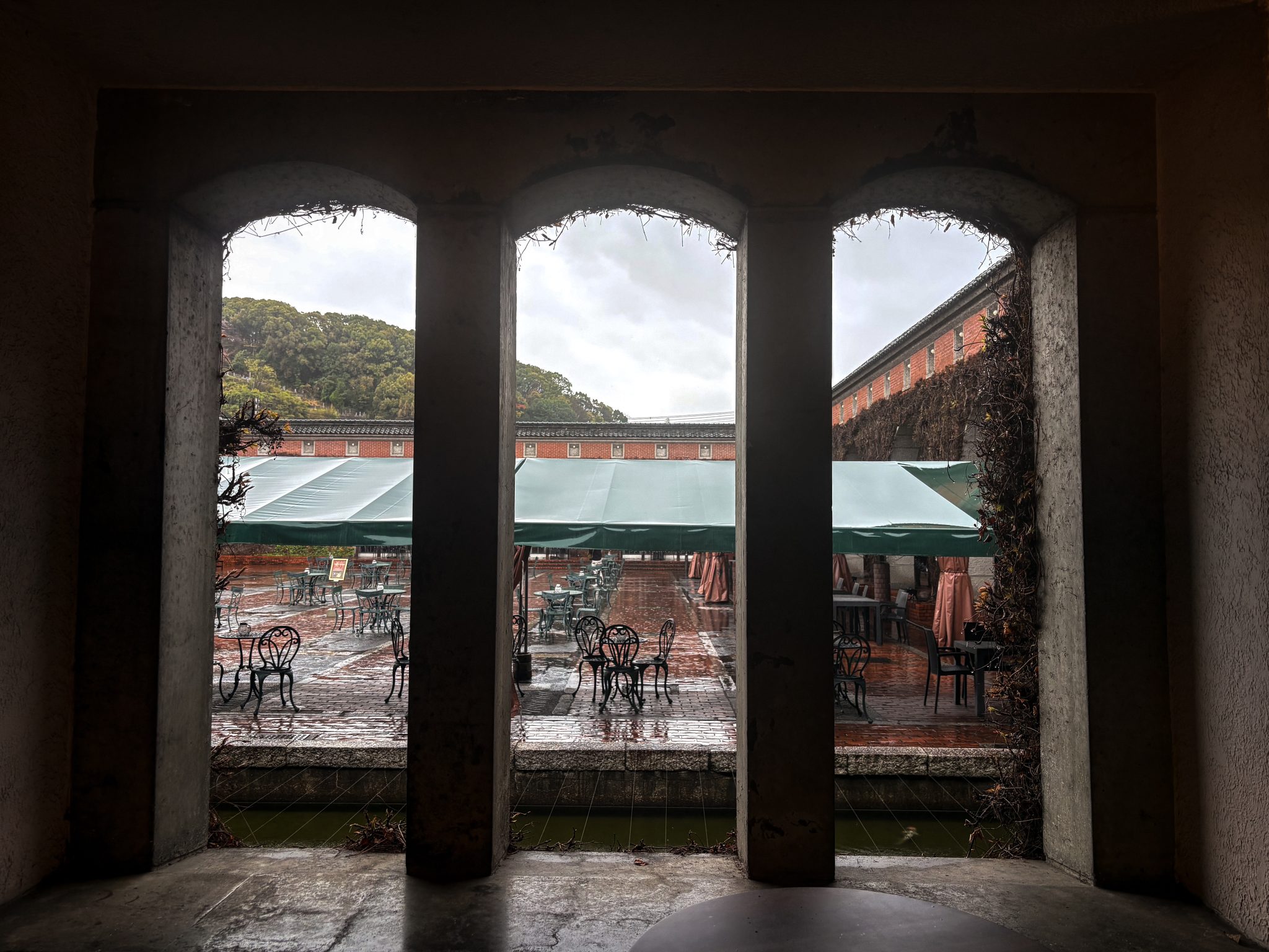 A view through three arched windows showing a rainy outdoor seating area. Outside are metal tables a